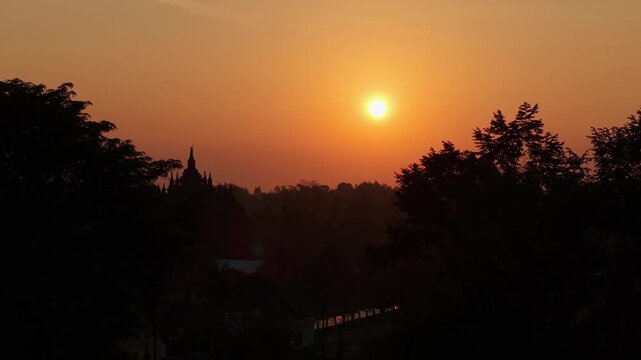 Aerial view of the silhouetted Plaosan Lor Temple during a golden sunset with orange sky and surrounding trees in Kecamatan Prambanan, Jawa Tengah, Indonesia.