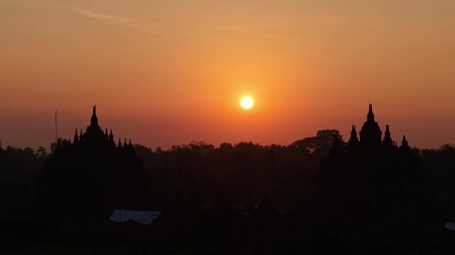 Aerial view of the silhouetted Plaosan Lor Temple during a vibrant orange sunset with the sun disk visible over the horizon in Kecamatan Prambanan, Jawa Tengah, Indonesia.