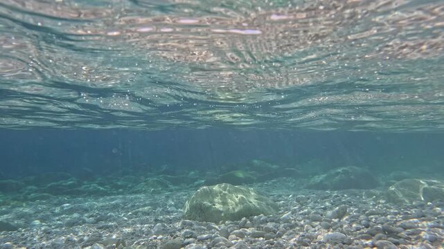 Underwater river flowing on pebbles and rocks. Crystal-clear water flows over smooth pebbles and rocks, creating a mesmerizing underwater scene with sunlight filtering through the surface