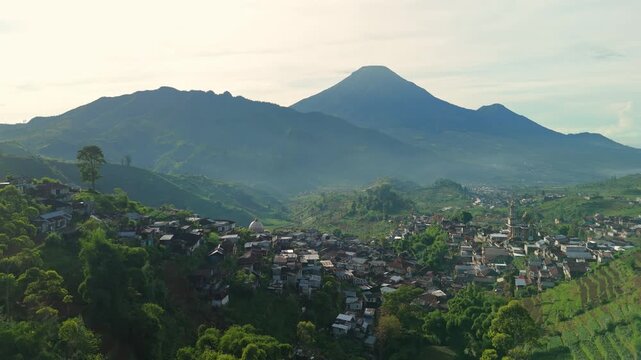 Aerial view of Dieng Plateau featuring a hillside village, terraced agricultural fields, and lush volcanic mountains under a clear sky in Kecamatan Batur, Jawa Tengah, Indonesia.