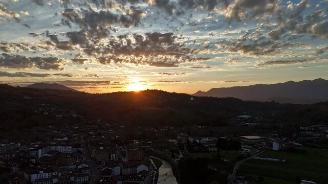 Aerial hyperlapse over Cangas de On&iacute;s at sunset, Asturias, Spain. Golden hour light and dramatic altocumulus clouds in orange and gold, with the Sella river winding through town below. DJI Mini 4 Pro,