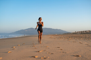 Obraz premium Barefoot woman jogging along wide sandy beach at sunrise, mountain backdrop, long trail of footprints, healthy outdoor workout, serene coastal landscape