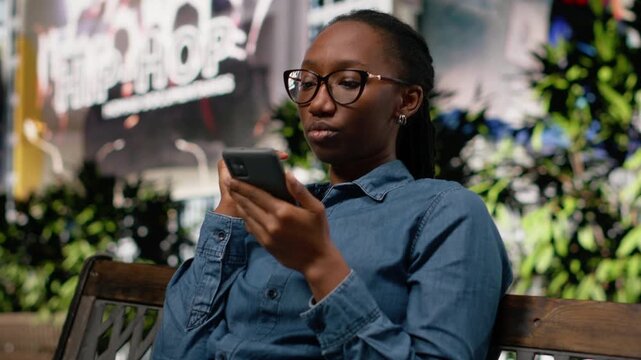 Black girl scrolling smartphone apps and relaxing on park bench, searching for map directions and things to do in the city. Pedestrian checking social media on phone, metropolitan street lifestyle.