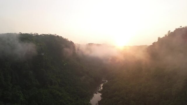 Aerial view of a misty valley with a winding river and lush green hills under a bright sunrise in Sukabumi, West Java, Indonesia.