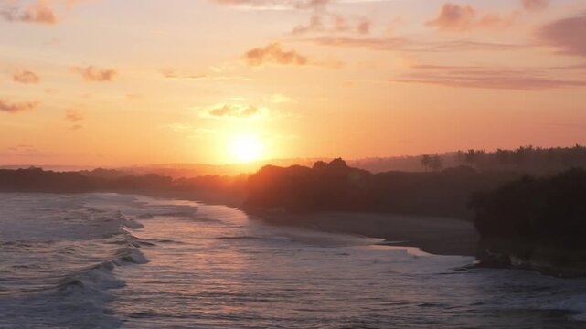 Aerial view of Ujung Genteng Beach at sunset with waves crashing on the shore and silhouettes of palm trees under a golden sky in Kecamatan Ciracap, Jawa Barat, Indonesia.