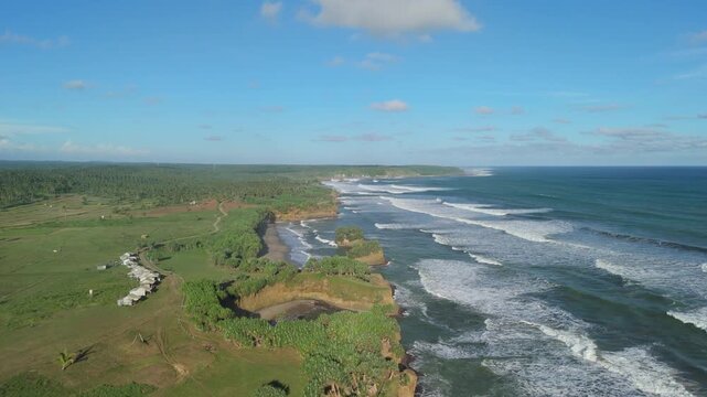 Aerial view of Ujung Genteng Beach with waves crashing against the rocky coastline and lush green fields under a blue sky in Kecamatan Ciracap, Jawa Barat, Indonesia.