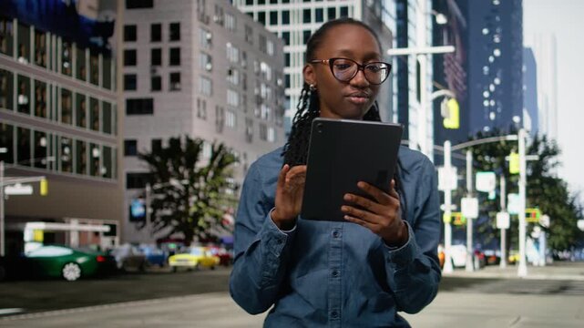 Smiling african american woman using tablet and walking in the city center, relaxing by looking at social media posts. Upbeat girl browsing navigation map in an urban landscape background.