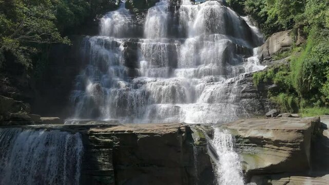 Aerial view of Cigangsa Waterfall with cascading water over tiered rock formations surrounded by lush green trees in Sukabumi, West Java, Indonesia.