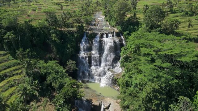 Aerial view of the multi-tiered Cigangsa Waterfall surrounded by lush tropical forest and terraced rice fields in Sukabumi, West Java, Indonesia.