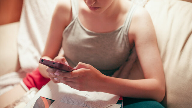 Focused high school girl sitting on a couch and typing on her mobile phone, using a smartphone to copy notes, browse exam answers, or cheat on a future test while studying at home.
