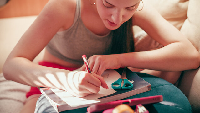 Close up of a concentrated teenage girl copying small text from her phone to create a tiny cheat sheet, preparing for an academic exam while sitting on a comfortable sofa in a living room.