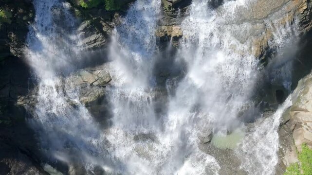 Aerial view of Cigangsa Waterfall cascading over dark rocky cliffs with white water mist and green foliage in the sunlight Sukabumi, West Java, Indonesia.