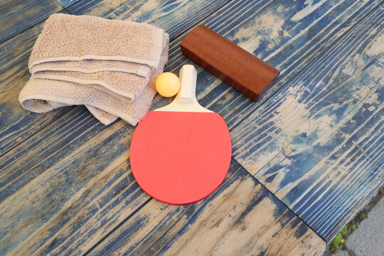Ping pong paddle rests beside towel and ball. Wooden table shows weathered blue paint. Equipment suggests casual game or sports hobby. No people visible, so emotions unneeded