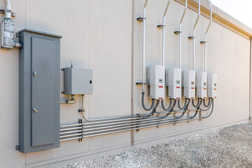 An outdoor installation of a commercial solar power system featuring a row of five wall-mounted grid-tie inverters connected via conduits to a large electrical distribution panel and disconnect box on © Andrei Liashchou