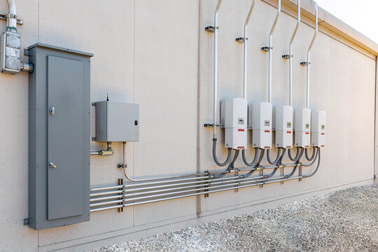 An outdoor installation of a commercial solar power system featuring a row of five wall-mounted grid-tie inverters connected via conduits to a large electrical distribution panel and disconnect box on