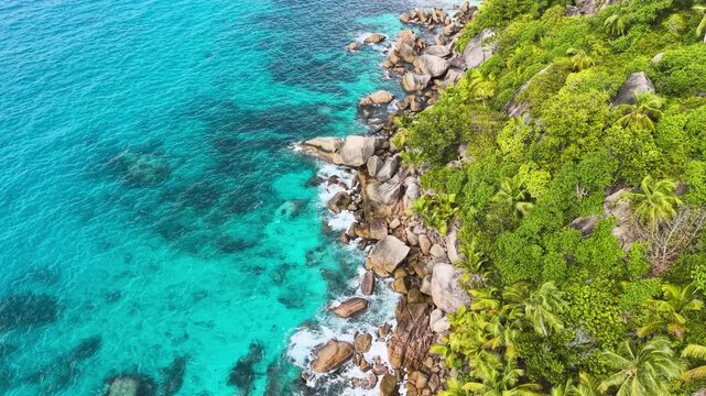 Aerial view of the turquoise ocean waves crashing against granite rocks and lush tropical palm trees on the shore of Felicite Island, La Digue, Seychelles.