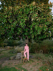 Fototapeta premium Young girl in pink overalls sits on a wooden chair beneath a lush tree in an open park, capturing a serene moment of solitude and nature, with soft light filtering through leaves.