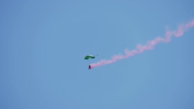 Skydiver descending with a colorful striped parachute canopy releasing a long pink smoke trail against a clear blue sky at an airshow