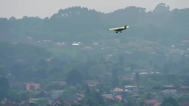 Small yellow high wing single engine light aircraft in level flight over hazy countryside with houses, trees and construction crane visible below