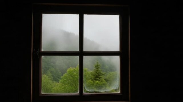 Moody view through a rustic wooden window frame looking out at a misty green forest landscape in the mountains