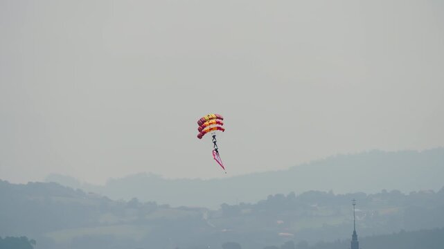 Skydiver under a red and yellow round parachute canopy descending through hazy sky carrying a flag with misty hills and coastline visible below