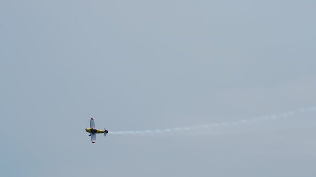 Brightly colored single-engine aerobatic monoplane in a steep banking turn leaving a wavy white smoke trail that curves across the gray sky