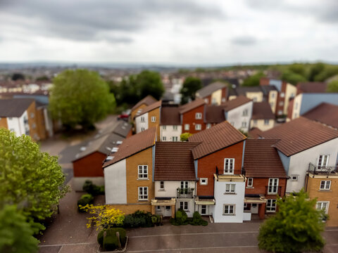A tilt-shift lens captures a miniature-like view of a neighborhood with colorful houses, green trees, and a cloudy sky, creating a whimsical and dreamy atmosphere.