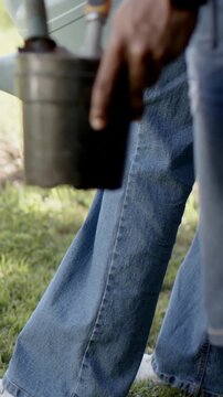 Vertical video: Aligning spout, two adults in jeans pouring water onto soil with blue can and tin