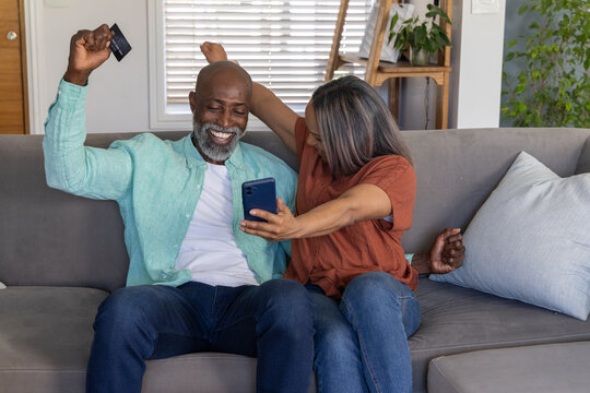 Senior couple sitting on gray sofa in living room, leaning holding smartphone and credit card