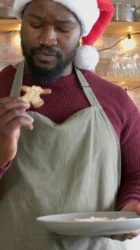 Vertical video: Picking up cookie, baker in olive apron sniffing, tasting in kitchen with Santa hat