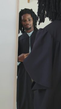Vertical video: Seeing mirror, man with dreads fastening dark gown at home, preparing for ceremony