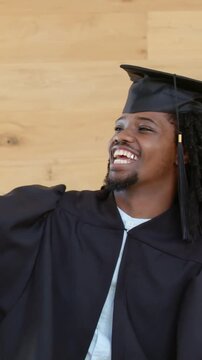 Vertical video: Extending arm, grad in gown taking phone selfie at wood wall, marking grad, tassel