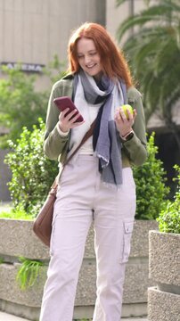 Vertical video: Walking woman after phone ping, scrolling and biting apple at plaza in olive jacket