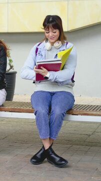 Vertical video: Smiling right, woman in denim paging yellow folder and red notebook at campus bench