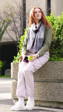 Vertical video: Sitting woman in jacket eating green apple and checking phone after buzz at plaza