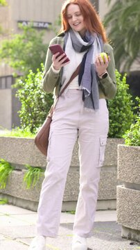 Vertical video: Glancing woman walking plaza in green jacket, smiling at pink phone, bringing apple