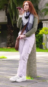 Vertical video: Leaning redhead woman in olive jacket raising phone to mouth at plaza, recording