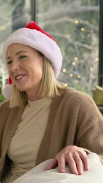 Vertical video: Laughing at offscreen cue, woman in Santa hat leaning on couch near tree, gesturing