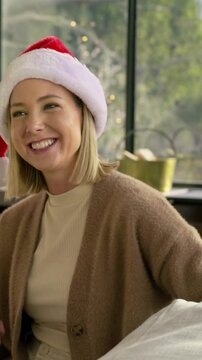 Vertical video: Receiving green gift, woman in Santa hat laughing, unwrapping on sofa for holiday