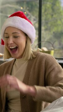 Vertical video: Smiling at offscreen friend, woman at home Santa hat giving green gift, copy space