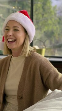 Vertical video: Hearing offscreen laugh woman in Santa hat, cardigan laughing and leaning at home
