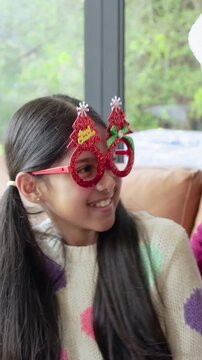 Vertical video: Leaning mother in Santa hat hugging daughter on couch at home, showing red glasses