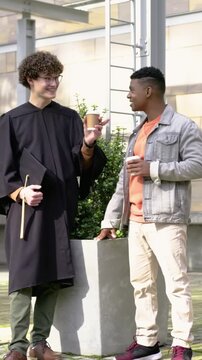 Vertical video: Raising paper cup, grad wearing gown and holding cap cheering with friend on campus