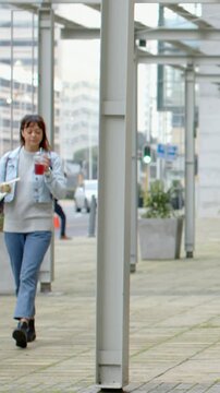 Vertical video: Approaching woman in denim under canopy holding food sipping drink hidden by column