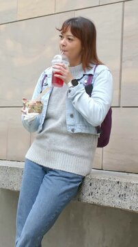 Vertical video: Settling woman sipping red cup at concrete ledge, eating takeout, watching street
