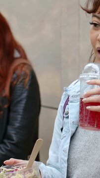 Vertical video: Glancing left, denim woman fixing straw, sipping, holding slaw at plaza with friend