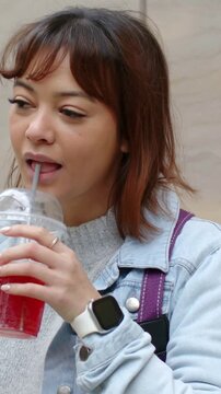 Vertical video: Looking down woman in denim lifting cup with straw tasting drink at stone wall