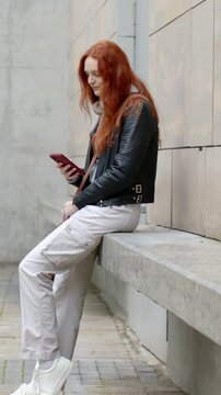 Vertical video: Holding pink cell woman reading texts on plaza bench in leather jacket, copy space
