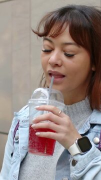 Vertical video: Moving cup into view, woman in denim holding cup and straw, sipping near stone wall