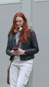 Vertical video: Scrolling redhead in jacket checking pink phone by wall as other passing, glancing